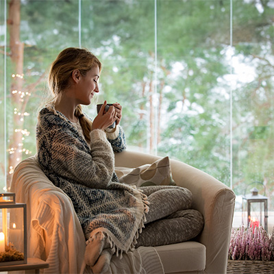 Frau sitzt eingekuschelt mit Tasse im Sessel vor Glasfront mit Winterblick – sanftes Licht, ruhige und gemütliche Atmosphäre