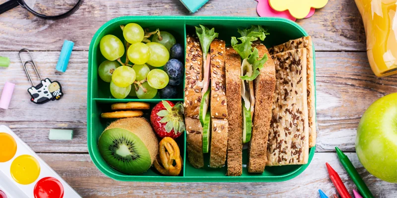 Lunchbox mit belegten Broten, Trauben, Kiwi, Erdbeere und Snacks neben bunten Schulsachen
