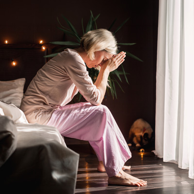 Ältere Frau sitzt nachdenklich am Bett im warmen Abendlicht – ruhige, intime Atmosphäre