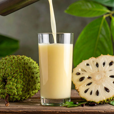 Cremiger Graviola-Saft wird ins Glas gegossen, daneben ganze und halbierte Frucht mit Kernen