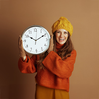 Lachende Frau in Herbstkleidung hält große Uhr vor braunem Hintergrund – warme Studiobeleuchtung, Symbol für Zeitumstellung