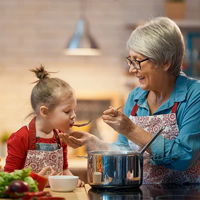 Ältere Frau reicht einem Kind beim Kochen einen Löffel zum Probieren – warme Küchenszene