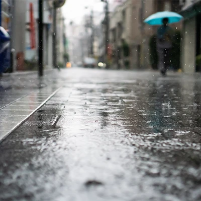 Regennasse Straße mit Spiegelungen und unscharfer Person mit türkisem Regenschirm im Hintergrund