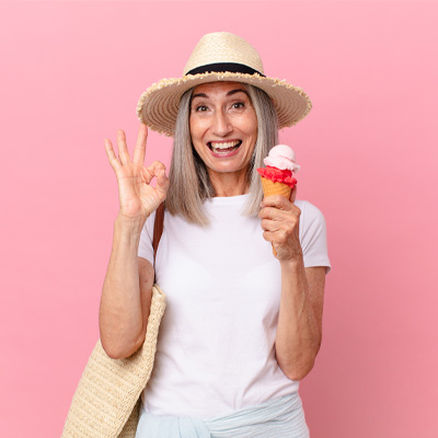 Fröhliche Frau mit Strohhut hält Eiswaffel und zeigt Okay-Geste vor rosa Hintergrund – helles Studiolicht, sommerliche Leichtigkeit