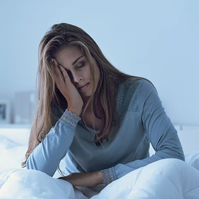 Tired-looking woman sitting in bed with her head resting on one hand – cool, subdued morning light
