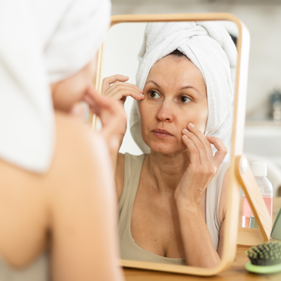 Frau mit Handtuch um den Kopf betrachtet ihre Haut im Spiegel – weiches Badezimmerlicht, ruhige Morgenroutine