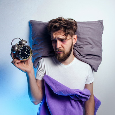 Mann liegt wach im Bett und hält einen Wecker in der Hand – kühles Licht, nächtliche und unruhige Stimmung