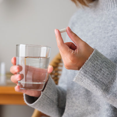 Person hält Glas Wasser und zeigt Tablette zwischen den Fingern – weiches Innenlicht, ruhige Alltagsszene