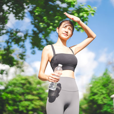 Frau in Sportkleidung hält Wasserflasche und schützt ihre Augen vor der Sonne – heller Sommertag