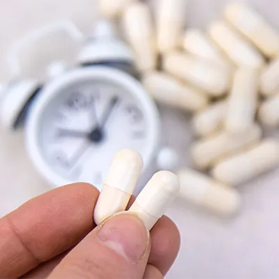 Hand hält zwei helle Kapseln vor weiteren Kapseln und einem unscharfen Wecker