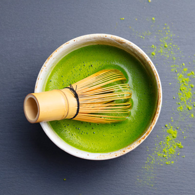Bowl of green matcha tea with bamboo whisk on dark surface – even lighting, minimalist presentation