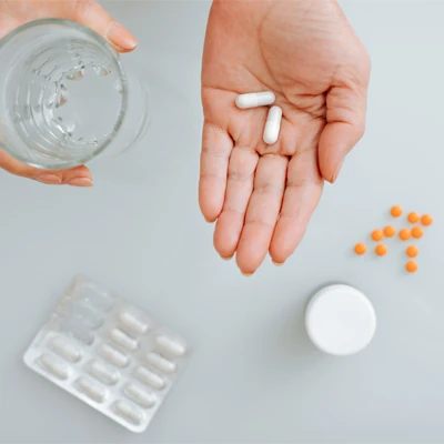 Hand hält zwei weiße Tabletten neben einem Glas Wasser und weiteren Medikamenten