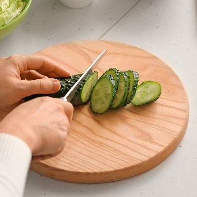 Hand slicing fresh cucumber on round wooden board – healthy preparation in bright kitchen
