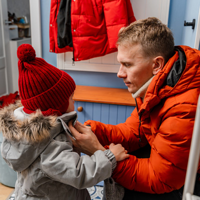 Vater zieht seinem Kind im Flur die Winterjacke an – warmes Licht, fürsorgliche Alltagsszene in der kalten Jahreszeit