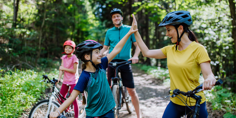 Family with bicycles on a forest trail, mother and child high-five – sunny daylight, active outdoor recreation
