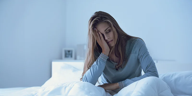 Tired-looking woman sitting in bed with her head resting on one hand – cool, subdued morning light