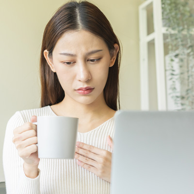 Woman holding a mug in front of a laptop, looking thoughtful – soft daylight, calm everyday scene