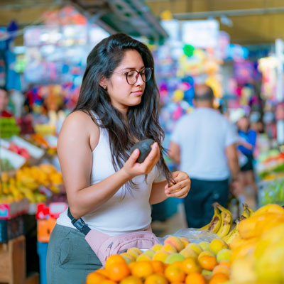 Frau prüft eine Avocado an einem Marktstand mit frischem Obst – lebendige Marktatmosphäre
