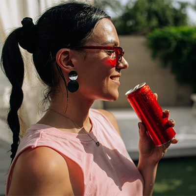 Smiling woman holding a red drink can in sunlight – summery scene with guarana context