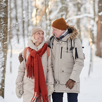 Älteres Paar in Winterkleidung spaziert lachend durch verschneiten Wald – helles Tageslicht, frische und lebendige Winterstimmung