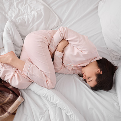 Frau liegt zusammengerollt im Bett – weiches Tageslicht, ruhige Innenaufnahme