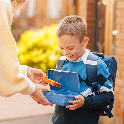 Junge mit Schulranzen freut sich über belegtes Brot in Brotdose – warmes Morgenlicht, Alltagsszene vor dem Schulstart