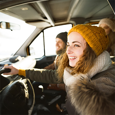 Junge Frau mit Wollmütze fährt fröhlich einen Van durch winterliche Landschaft – helles Sonnenlicht, lebendige Reiseszene