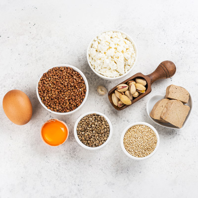 Proteinreiche Lebensmittel wie Ei, Hüttenkäse, Samen, Getreide und Tofu auf hellem Untergrund