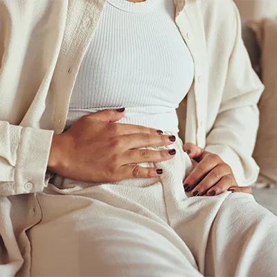 Seated woman in light-colored clothing resting one hand on her abdomen – soft daylight, calm atmosphere