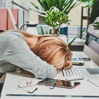 Erschöpfte Frau liegt mit Kopf auf dem Schreibtisch neben Computer und Smartphone – helles Bürolicht