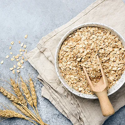 Bowl of rolled oats with wooden scoop on linen cloth, oat stalks beside it – natural source of beta-glucan
