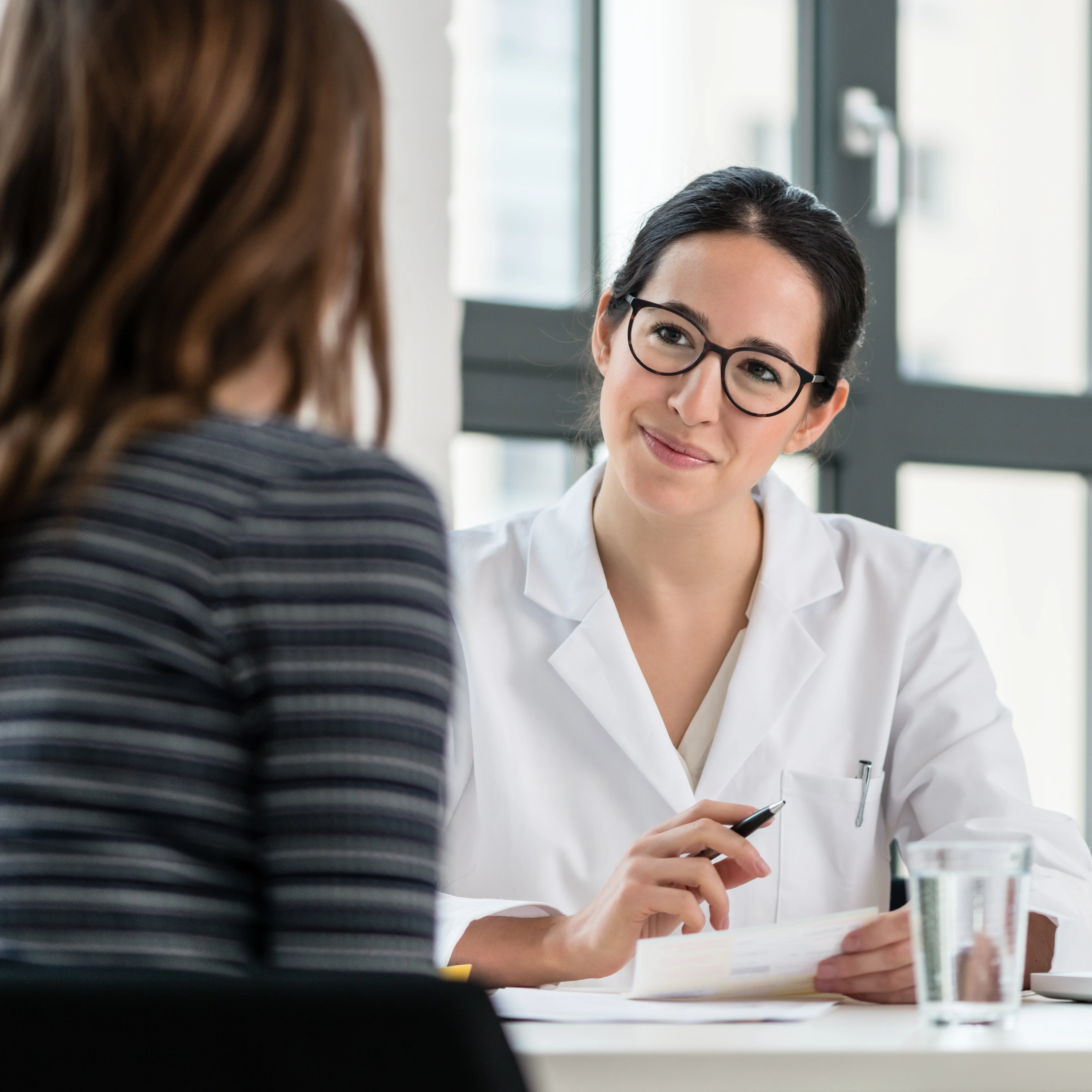 Ärztin mit Brille und weißem Kittel spricht mit Patientin in heller Praxis – freundliche Gesprächsatmosphäre