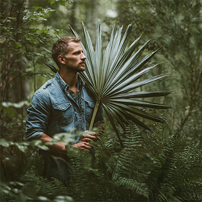 Mann mit Bart steht in dichter grüner Natur und hält großes Palmenblatt – weiches Umgebungslicht, ruhige Waldstimmung