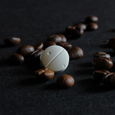 White caffeine tablet among coffee beans on dark fabric – high-contrast close-up